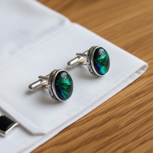 May include: A pair of silver cufflinks with oval-shaped, iridescent green and blue stones. The cufflinks are set in a decorative silver frame and are displayed on a white dress shirt cuff, with a wooden surface in the background.