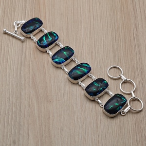 May include: A silver bracelet with six rectangular black opal stones displaying green and blue iridescence. The bracelet has a toggle clasp and a double-ring clasp. The jewellery is presented on a light wood surface.