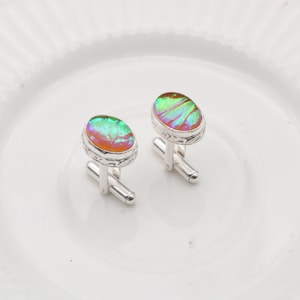 Op de afbeelding: Een paar zilveren manchetknopen met ovale, iriserende stenen. De stenen vertonen een mix van groene, oranje en paarse tinten. De manchetknopen liggen op een witte plaat.