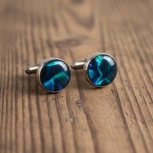 May include: A pair of round cufflinks with a silver-colored metal base. The center of each cufflink features a swirling pattern of blue and green colors. The cufflinks are displayed on a wooden surface.