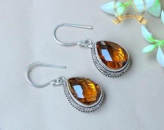 May include: A pair of silver-toned drop earrings. Each earring features a faceted, teardrop-shaped amber-colored gemstone set in a detailed, patterned bezel. The earrings have a simple hook closure.