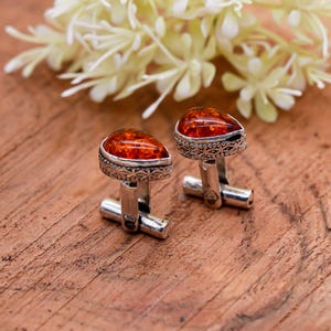May include: Silver cufflinks featuring amber-coloured, teardrop-shaped stones. The cufflinks have a detailed, patterned border and a classic T-bar closure. The background includes white flowers and a wooden surface.