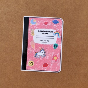 May include: Pink composition book with a black spine and colorful stickers. The stickers include a unicorn, a rainbow, a peace sign, a heart, a smiley face, a UFO, an ice cream cone, a flower, and a red lip. The book says "Composition Book" and "100 sheets 8 1/2 x 11"