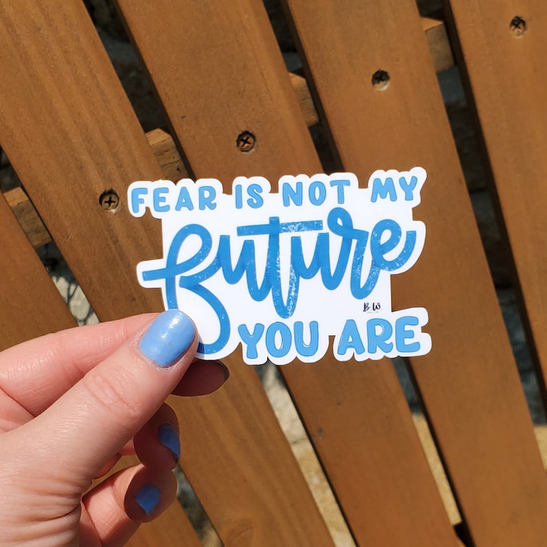 May include: A blue and white sticker with the text "Fear is not my future you are" in a handwritten font. The sticker is being held by a hand with blue nail polish.
