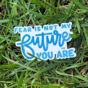 May include: A blue and white sticker with the text "Fear is not my future you are" in a handwritten style. The sticker is on a bed of green grass.