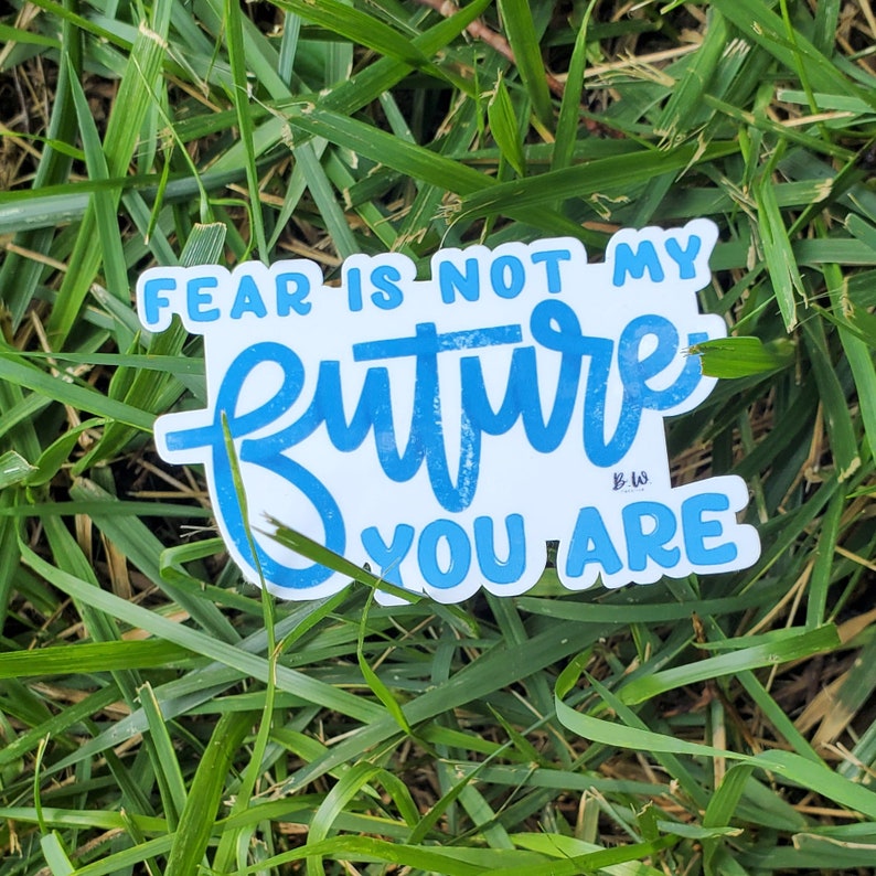 May include: A blue and white sticker with the text "Fear is not my future you are" in a handwritten style. The sticker is on a bed of green grass.