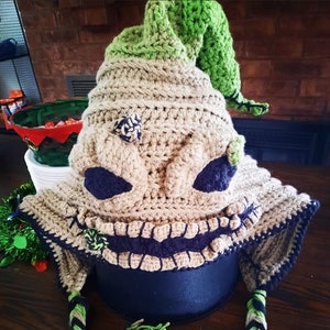 Pode incluir: Chapéu de crochê em um esquema de cores verde e marrom, com um personagem com um grande sorriso cheio de dentes. O chapéu tem um cordão longo trançado com detalhes verdes e marrons.