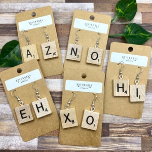 May include: A set of eight pairs of earrings featuring wooden Scrabble tiles with letters and numbers. The tiles are attached to silver earring hooks. The earrings are displayed on brown cardstock with the name "ROVEWORKS STUDIO" printed on each card.