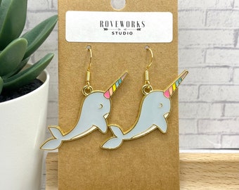 May include: A pair of gold tone earrings featuring a narwhal design. The narwhals are light blue with a rainbow colored horn. The earrings are on a brown card with the text "ROVEWORKS STUDIO".