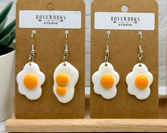 May include: Two pairs of dangling earrings shaped like fried eggs, attached to brown cardboard display cards labelled Roveworks Studio. Each earring features a white egg white shape with a glossy, raised, bright orange yolk centre.