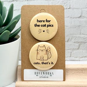 May include: Two circular cat-themed badges on a brown card. The top badge reads "here for the cat pics" with a cat face graphic. The bottom badge says "cats. that's it." with a cat illustration. The card has a white label with "ROVEWORKS STUDIO" text.