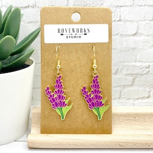 Peut inclure: Une paire de boucles d'oreilles dorées avec des breloques de fleurs violettes. Les boucles d'oreilles sont sur une carte brune avec le texte "ROVEWORKS STUDIO".