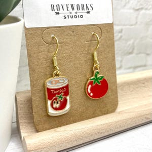 May include: A pair of gold dangle earrings with a red tomato and a can of tomato soup charm. The charms are enamel and have a white background with black outlines.