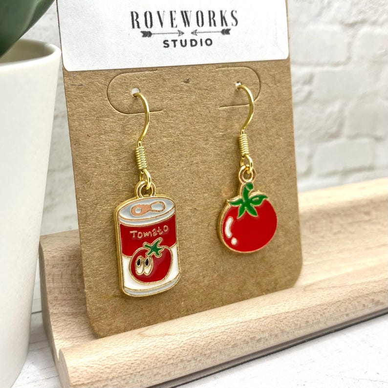 May include: A pair of gold dangle earrings with a red tomato and a can of tomato soup charm. The charms are enamel and have a white background with black outlines.