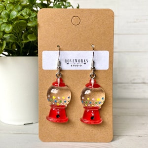 May include: A pair of red gumball machine earrings with silver hooks. The gumball machines are clear and filled with colourful gumballs.