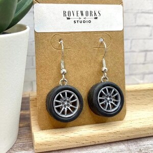 May include: A pair of novelty earrings shaped like miniature black car tyres with silver rims. The earrings hang from silver hooks and are displayed on a brown card with the text "ROVEWORKS STUDIO".