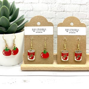 May include: Two pairs of dangle earrings featuring a red tomato and a can of tomatoes. The earrings are on brown card stock with the text "ROVEWORKS STUDIO" printed on each card.