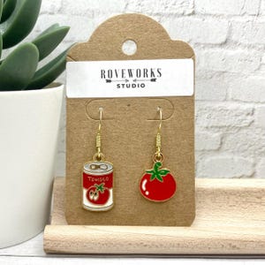 May include: Gold dangle earrings with a red tomato and a red can of tomato soup. The can has the word "Tomato" on it. The earrings are on a brown card with the text "Roveworks Studio".