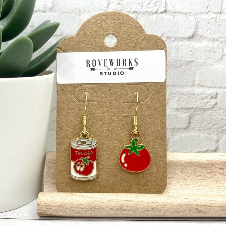 May include: Gold dangle earrings with a red tomato and a red can of tomato soup. The can has the word "Tomato" on it. The earrings are on a brown card with the text "Roveworks Studio".