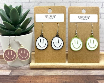 May include: Three pairs of smiley face earrings on display cards. The earrings are black, green, and pink with white outlines. The cards have the text "ROVEWORKS STUDIO" printed on them.