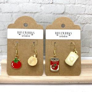 May include: Two pairs of gold dangle earrings with food-themed charms. The first pair features a red tomato and a golden wheel of pasta. The second pair features a gold can of tomatoes and a golden square of bread.