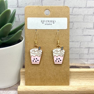 Peut inclure: Boucles d'oreilles pendantes dorées représentant un gobelet à boba en émail rose et blanc avec des perles de boba noires et une paille grise. Les boucles d'oreilles sont sur une carte brune avec le texte "Roneworks Studio".