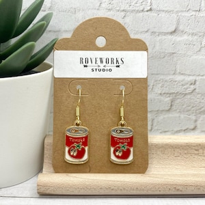 May include: Gold dangle earrings featuring a red tomato can charm with the word "Tomato" printed on the can. The earrings are displayed on a brown card with the text "Roveworks Studio".