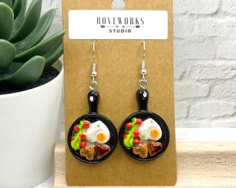 May include: A pair of novelty earrings featuring miniature black frying pans. Each pan holds a fried egg, bacon, and vegetables. The earrings hang from silver-colored hooks and are displayed on a brown card with the text "ROVEWORKS STUDIO".
