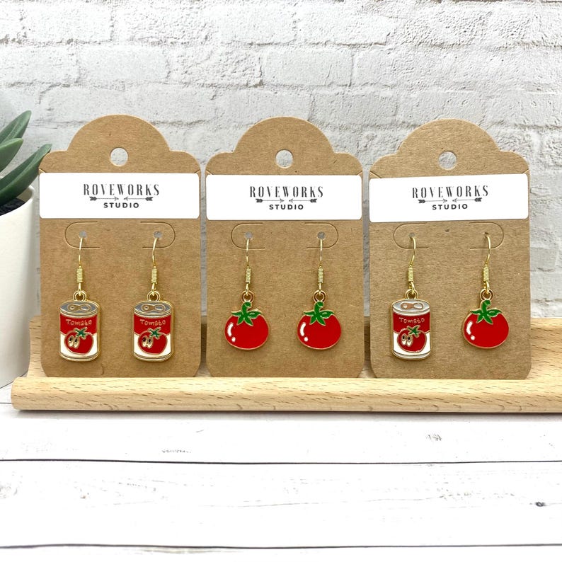 May include: Three pairs of gold dangle earrings with red tomato and tomato can charms. The earrings are displayed on brown card stock with the text "ROVEWORKS STUDIO" printed on each card.