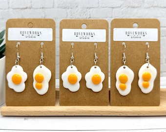 May include: Three pairs of dangling earrings shaped like fried eggs, each featuring a white egg white and a bright yellow yolk. The earrings are attached to brown cardboard display cards labeled Roveworks Studio with silver-toned hooks.