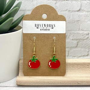 May include: Gold dangle earrings with red tomato charms. The earrings are on a brown card with the text "ROVEWORKS STUDIO".