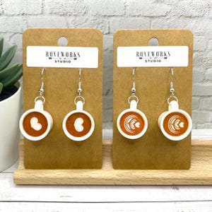 May include: A pair of coffee cup earrings featuring a latte art design. The earrings are white with a brown interior and silver hooks. They are displayed on a brown card with the text "ROVEWORKS STUDIO".