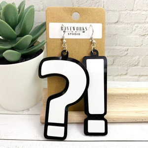May include: A pair of black and white earrings shaped like a question mark and an exclamation point. The earrings are on a brown card with the text "ROVEWORKS STUDIO".