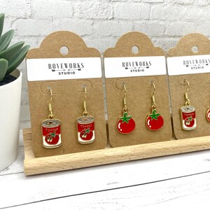 May include: Three pairs of gold dangle earrings on brown card stock. The earrings feature a red tomato and a can of tomatoes. The card stock has the text "ROVEWORKS STUDIO" printed on it.