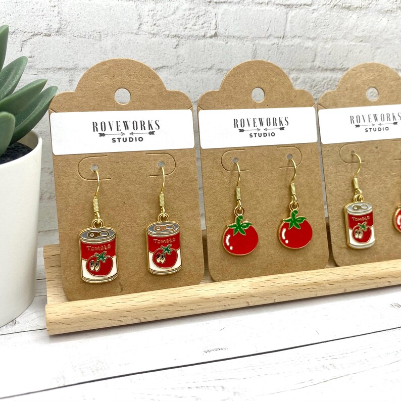 May include: Three pairs of gold dangle earrings on brown card stock. The earrings feature a red tomato and a can of tomatoes. The card stock has the text "ROVEWORKS STUDIO" printed on it.
