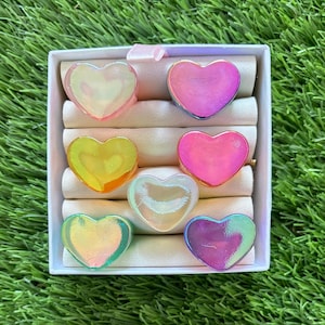 Puede incluir: Ocho anillos en forma de corazón iridiscentes en una caja blanca. Los anillos son de varios colores, incluyendo rosa, morado, azul, verde, amarillo y blanco.
