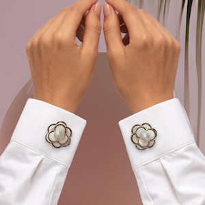 May include: White dress shirt with flower-shaped cufflinks. The cufflinks have a gold-colored outline and a white, pearl-like center. The shirt sleeves are neatly folded and buttoned.