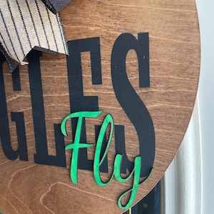 May include: A round wooden sign with the words "EAGLES Fly" in black and green. The sign has a dark brown stain and a black and tan striped bow at the top.