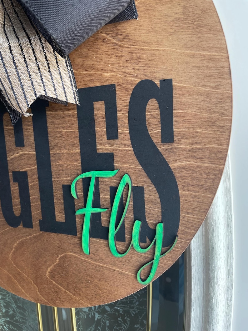 May include: A round wooden sign with the words "EAGLES Fly" in black and green. The sign has a dark brown stain and a black and tan striped bow at the top.