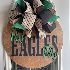 May include: A round wooden door sign with the words "EAGLES Fly Fly" in black and green. The sign is decorated with a large bow made of black, green, and tan striped ribbons.