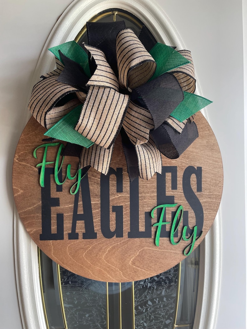 May include: A round wooden door sign with the words "EAGLES Fly Fly" in black and green. The sign is decorated with a large bow made of black, green, and tan striped ribbons.