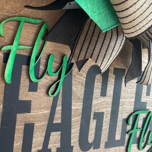 May include: Wooden sign with the words "Fly EAGLES Fly" in black and green. The sign has a wooden background with a decorative bow in green, black, and striped tan ribbon.