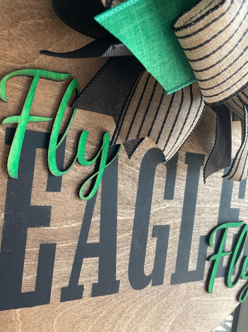 May include: Wooden sign with the words "Fly EAGLES Fly" in black and green. The sign has a wooden background with a decorative bow in green, black, and striped tan ribbon.