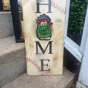 May include: A wooden sign with the word "HOME" in black capital letters. A green, red, and blue cartoon character is in the center. Baseball stitching accents the sign. The sign is weathered with a light brown background.