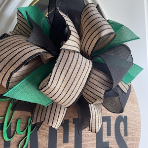 May include: A wooden round sign with the words "Fly Eagles Fly" in black and green lettering. The sign is decorated with a large bow made of black, green, and tan striped ribbons. The bow is positioned at the top of the sign.