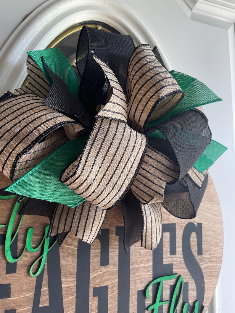 May include: A wooden round sign with the words "Fly Eagles Fly" in black and green lettering. The sign is decorated with a large bow made of black, green, and tan striped ribbons. The bow is positioned at the top of the sign.
