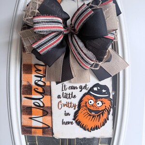 Op de afbeelding: Een decoratief deurbord met een grote zwarte strik met rood-wit gestreept lint. Het bord heeft een welkomstboodschap en een Gritty-karakterafbeelding met de tekst "It can get a little Gritty in here."