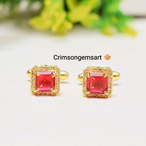 May include: A pair of gold cufflinks with a square red gemstone surrounded by smaller white gemstones.