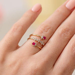 May include: A gold-coloured wrap ring with three spirals. The ring features small, round, red gemstones and clear, sparkling accents. The ring is displayed on a hand with a light skin tone, showcasing the jewellery's design and details.