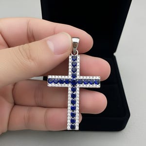 Pu&ograve; includere: Ciondolo a forma di croce in argento con un alone di piccole pietre bianche e pietre blu pi&ugrave; grandi. La croce &egrave; tenuta davanti a un portagioie nero.